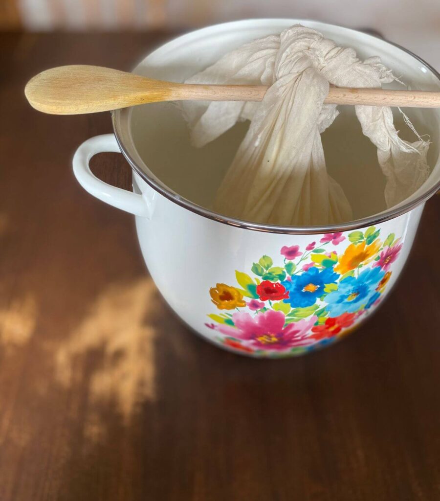 the-humming-homemaker-easy-homemade-instant-pot-cream-cheese-pioneer-woman A cheesecloth of yogurt tied to a wooden spoon hanging over a large decorative Pioneer Woman pot.