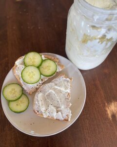 Bird's eye view of a white plate of sourdough toast smothered in homemade cream cheese topped with cucumber slices and pepper.