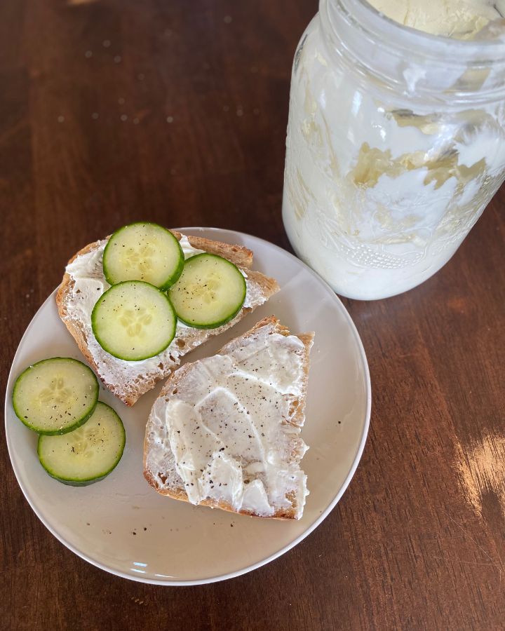 Bird's eye view of a white plate of sourdough toast smothered in homemade cream cheese topped with cucumber slices and pepper.