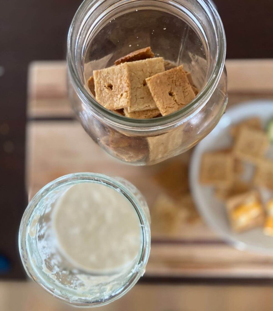 A bird's eye view of a messy glass jar of sourdough discard and a jar of crispy golden crackers. A snacking plate of crackers and cheese awaits. 