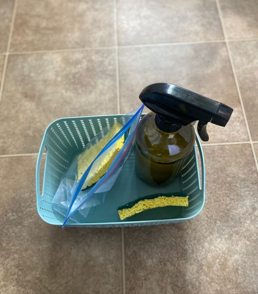 A plastic basket with a glass cleaner spray bottle, a dual-sided sponge and another dual-sided sponge in an open ziplock bag. 