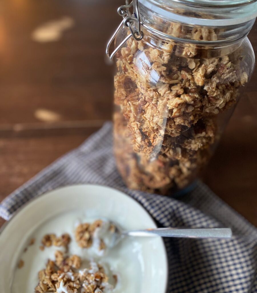 A large flip-top jar of homemade granola with a delicious-looking bowl of yogurt and granola with a spoonful ready to be eaten.
