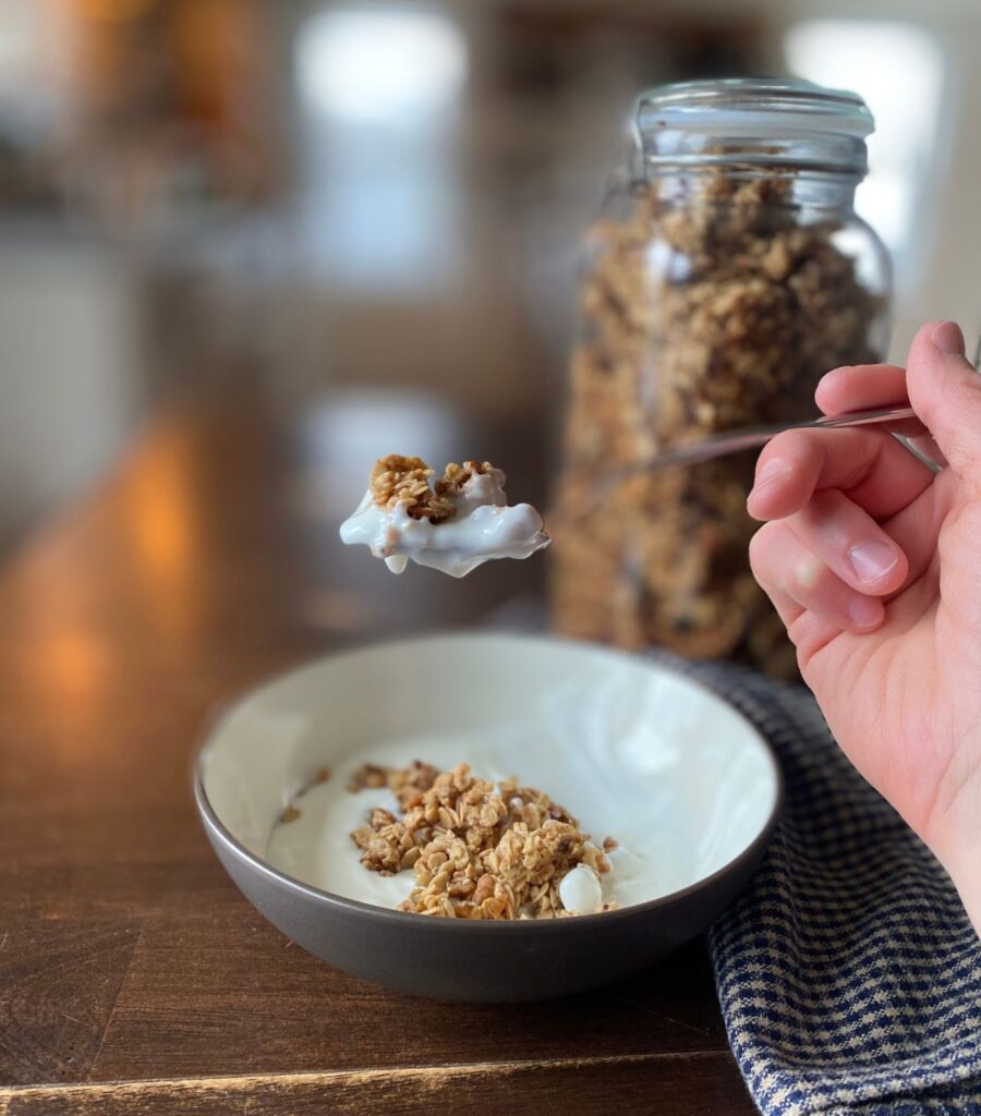 Someone holding a delicious-looking spoonful of creamy yogurt and crunchy homemade granola in front of a jar full of granola.