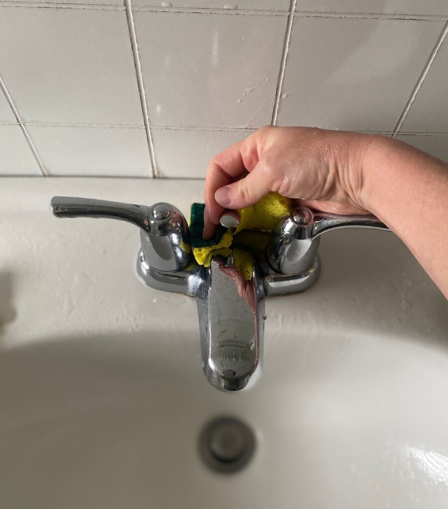 Scrubbing all the nooks and crannies of a chrome bathroom faucet with a green and yellow dual-sided sponge.