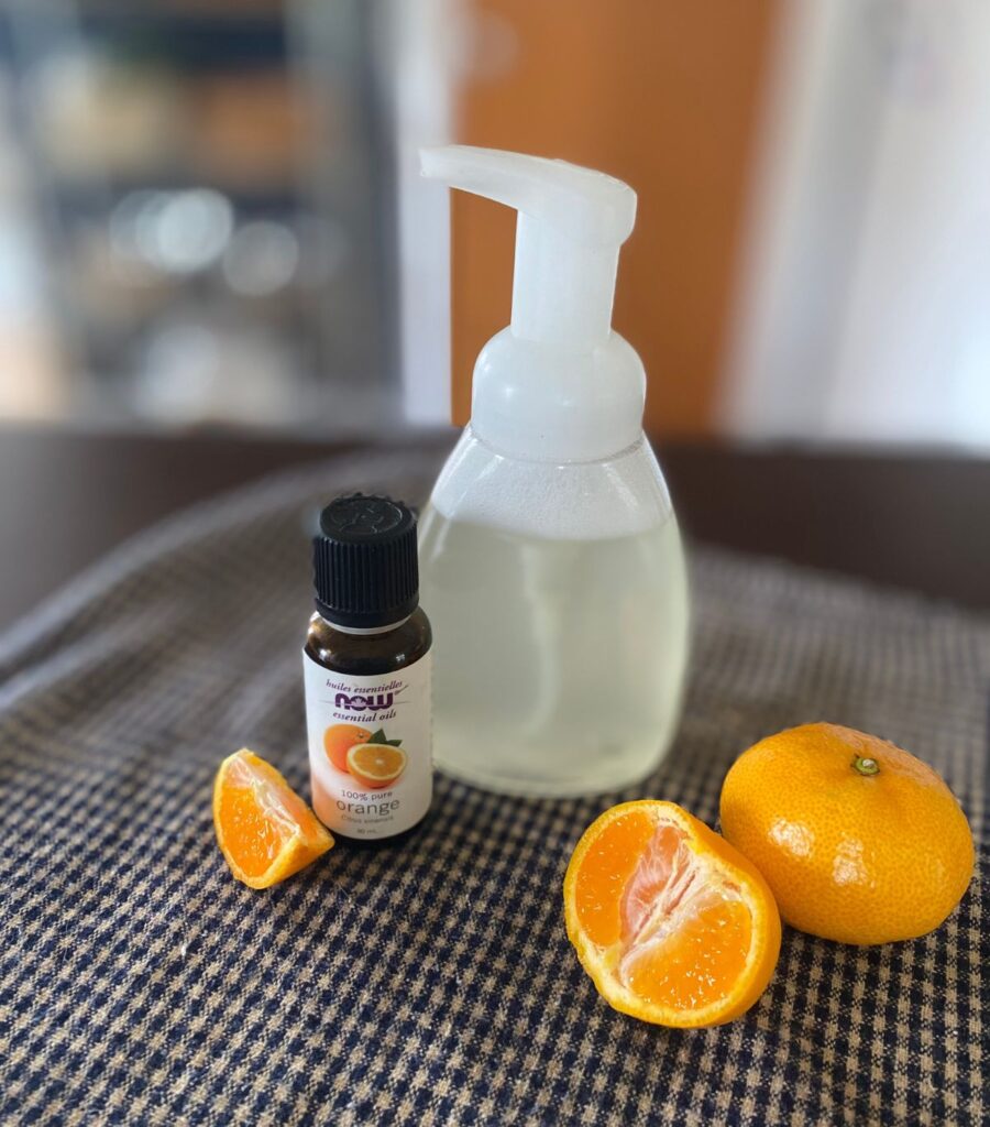 A clear dispenser of DIY foaming hand soap sitting on a table with sliced and halved clementines and orange essential oil.