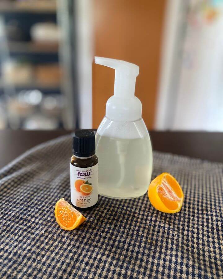 A clear foaming soap dispenser of DIY hand soap displayed on a table with orange essential oil and a halved and sliced clementine.