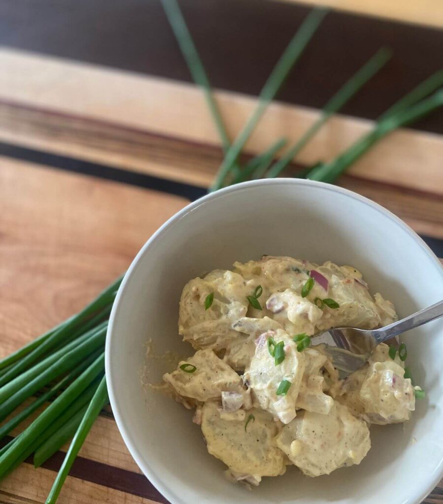 A delicious bowl of loaded potato salad topped with fresh chives sitting on a cutting board.