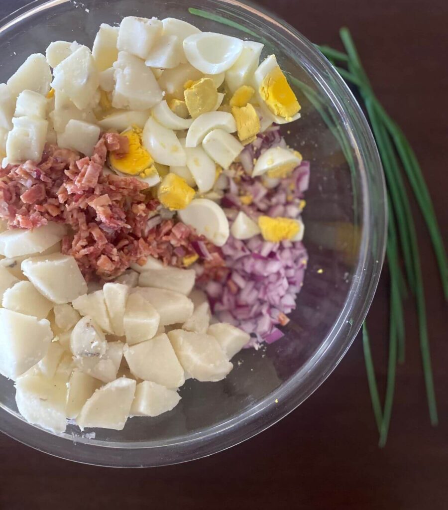 All the bright ingredients of loaded potato salad in glass bowl without the dressing with fresh chives on the table.
