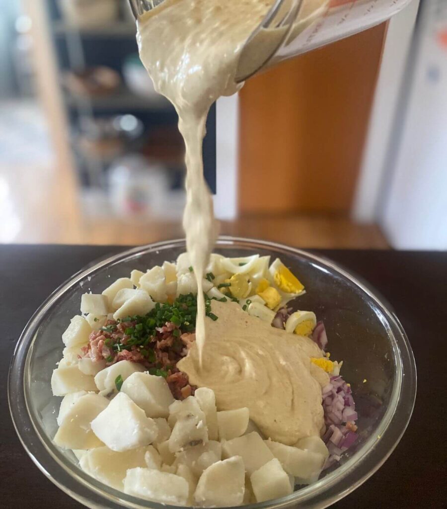 Pouring the creamy dressing over into the bowl to complete the loaded potato salad.