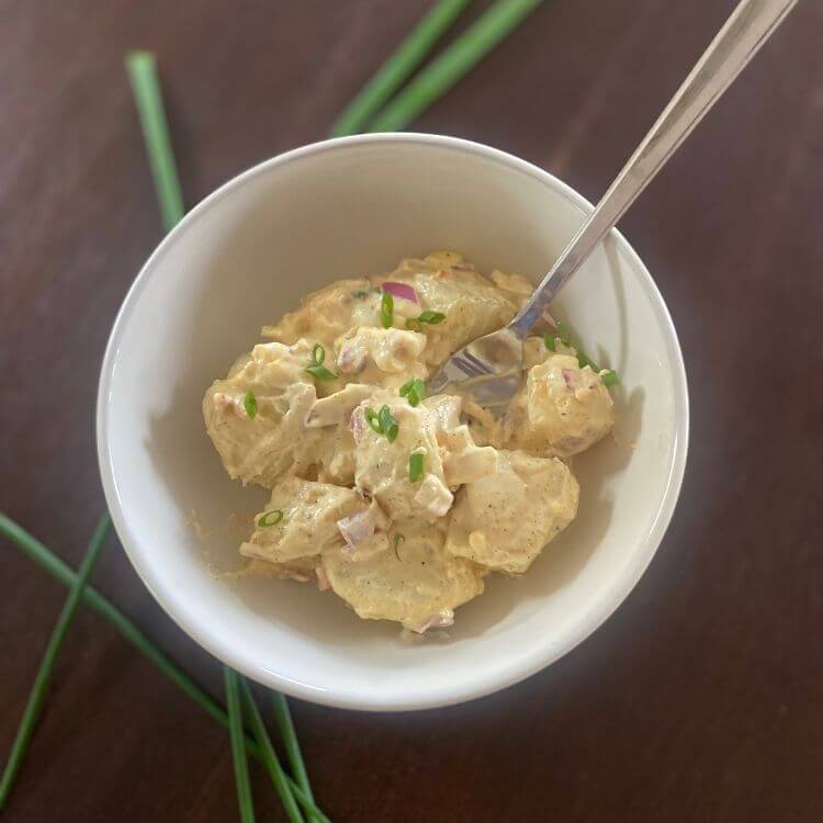 A bowl of creamy potato salad on a wooden table with fresh chives surrounding it.