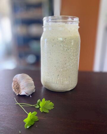 A large jar of fresh creamy homemade ranch dressing on a wooden table beside a head of garlic and fresh parsley.